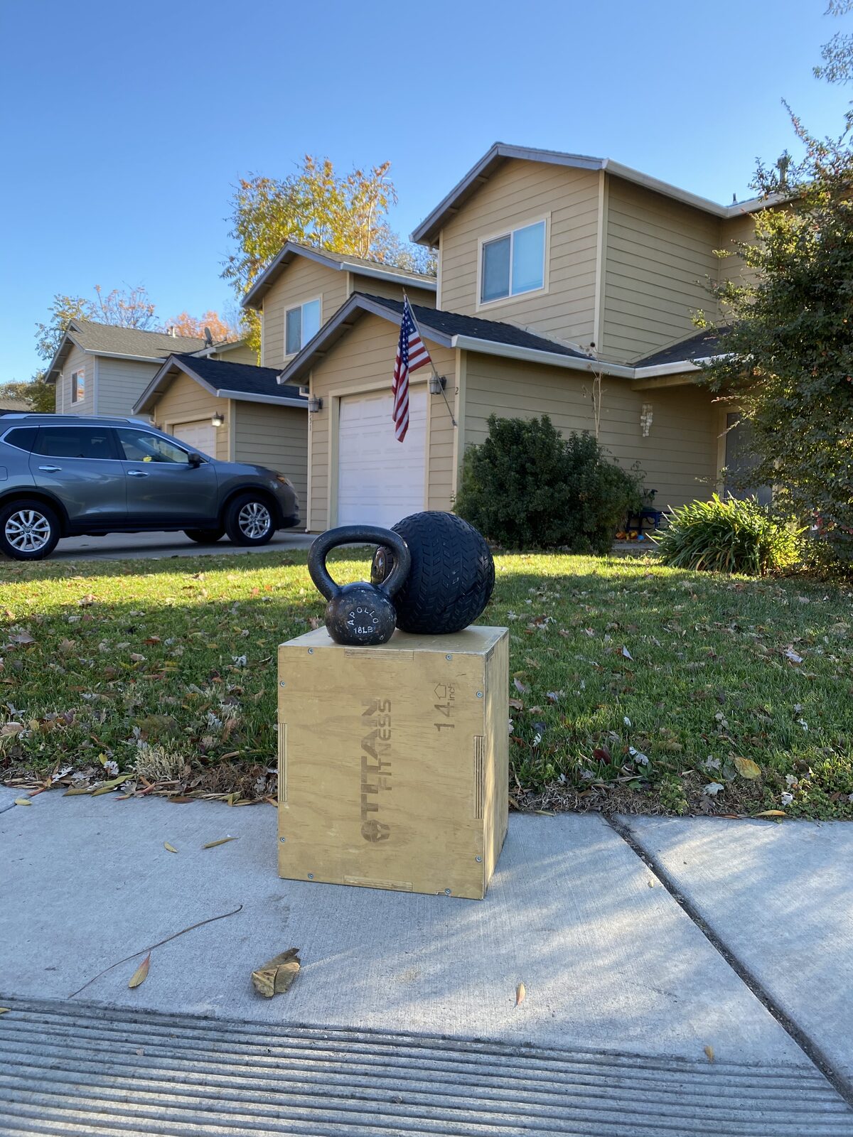 A kettlebell and a heavy tread-style medicine ball rest on a wooden plyo box in a sunny residential driveway in Chico, CA, showing the convenience of at-home personal training with Gym Truck Chico.